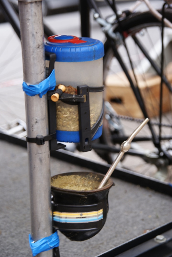 Clear plastic container filled with loose yerba mate above a gourd with a metal straw, all strapped to a metal frame.