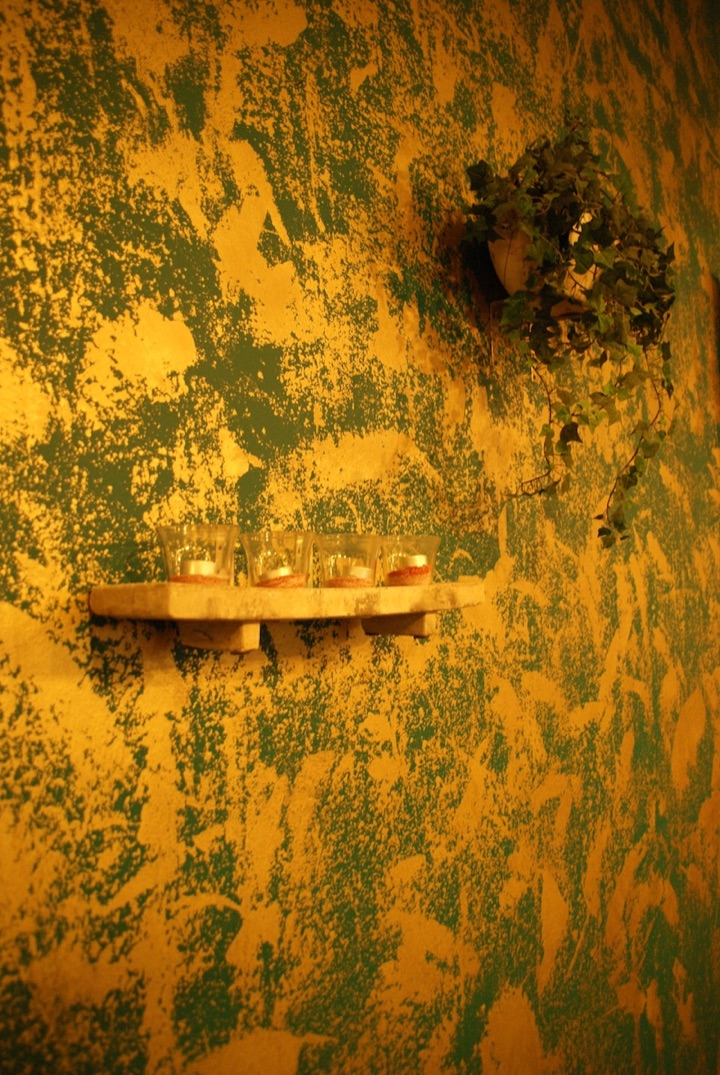 Glass candle holders on a small shelf against a textured yellow-and-green plaster wall, with a potted plant above.