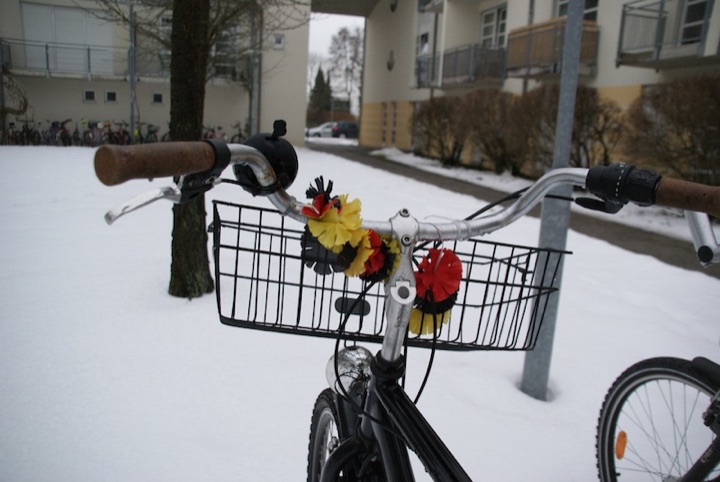 A bicycle with flowers in the black, red, and gold colors of the German flag on a snowy residential street.