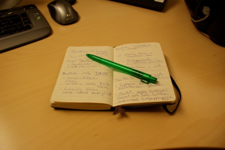 Open notebook with handwritten thesis notes and a green pen on a desk.