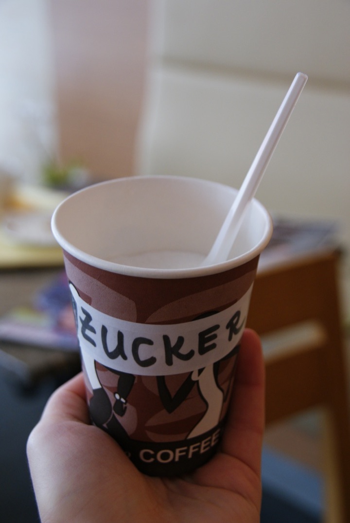 Hand holding a takeaway coffee cup labeled ‘ZUCKER’ with a white plastic spoon.