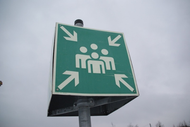 Green sign on a pole showing three people and arrows pointing toward them.