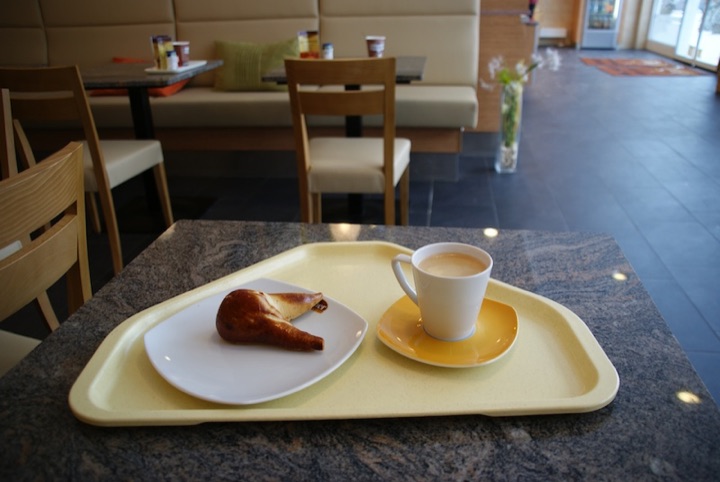 Yellow cafeteria tray holding a small cup of coffee and a partly eaten Bavarian Nussbeugerl pastry on a table.
