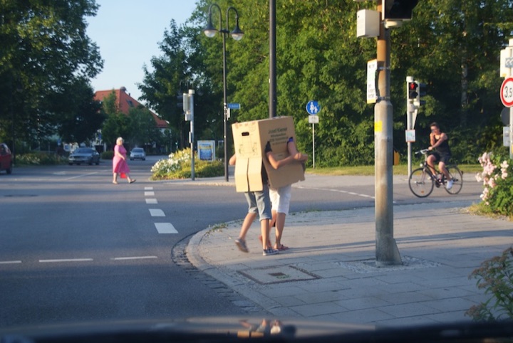 Two people walking inside a large cardboard box at a street crossing.