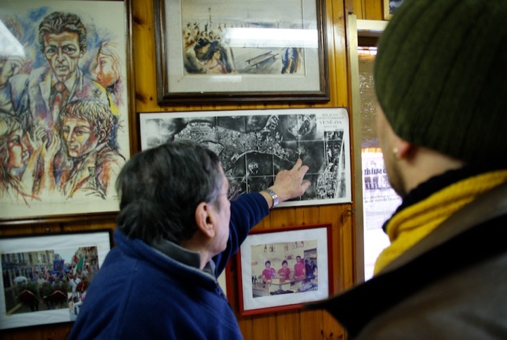 Man pointing at a framed aerial photograph on a wood-paneled wall while another person looks on.