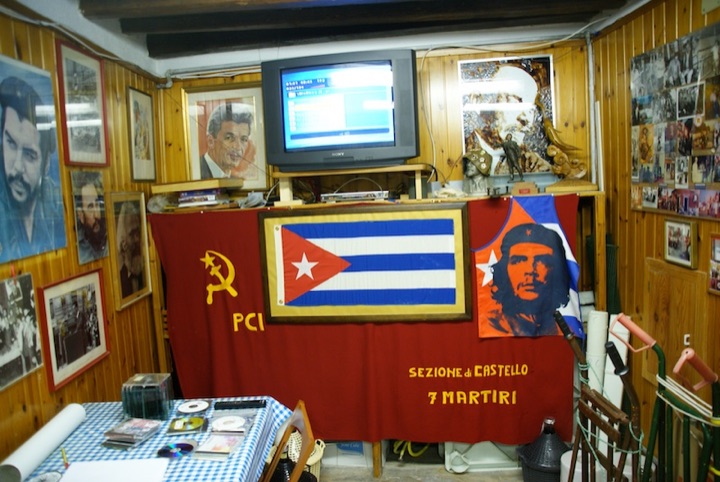 Wood-paneled room displaying framed portraits, a Cuban flag, and a red hammer-and-sickle banner beneath a television.