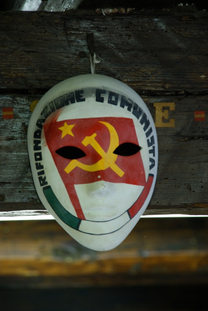 Painted white face mask with a red hammer-and-sickle emblem and Italian lettering against a dark background.