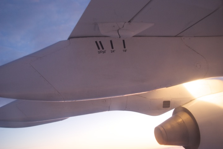 Airplane wing and engine viewed through a window with sunlight at the horizon.