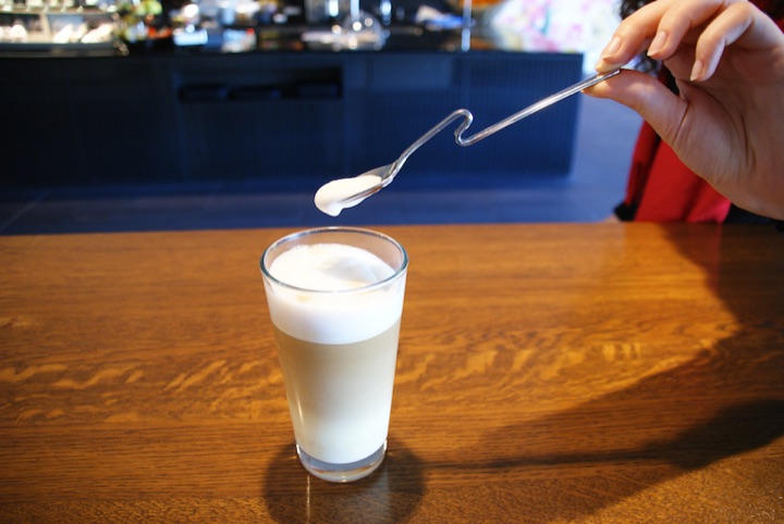 Hand holding a spoon above a glass of milk.