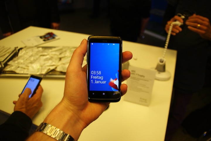 Hand holding a black HTC smartphone with a blue screen; another phone and people appear behind it.