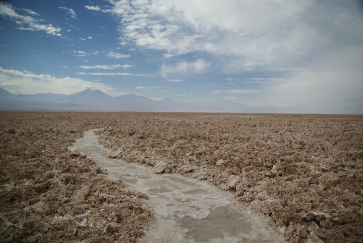 Rocky flat landscape with a shallow channel and distant mountains beneath a partly cloudy sky.