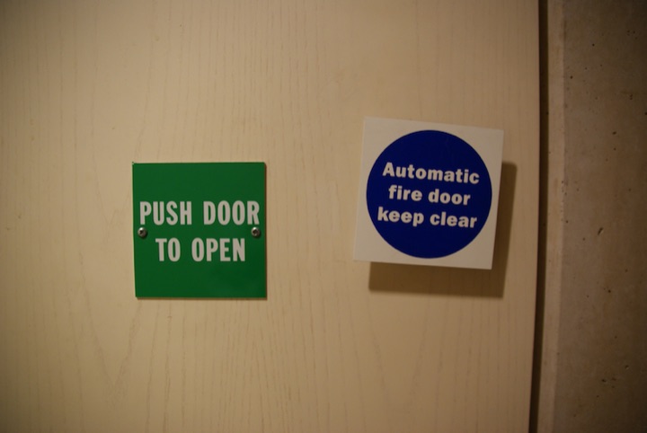 Door with green sign reading ‘PUSH DOOR TO OPEN’ beside blue sign reading ‘Automatic fire door keep clear.’