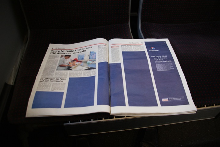 Open newspaper on a dark red-patterned train seat, with a blue Swisscom advertisement on the right page.