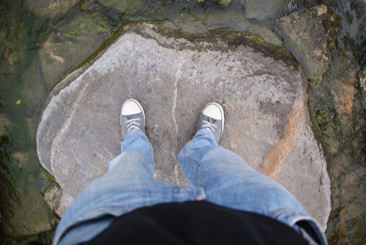 Person standing on a rock in shallow moving water, seen from above.