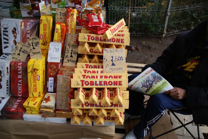Stacks of Toblerone and other chocolate bars at a street stall beside a seated person holding a newspaper.