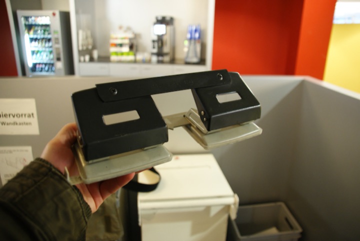 Hand holding two metal hole punches above a white office bin.