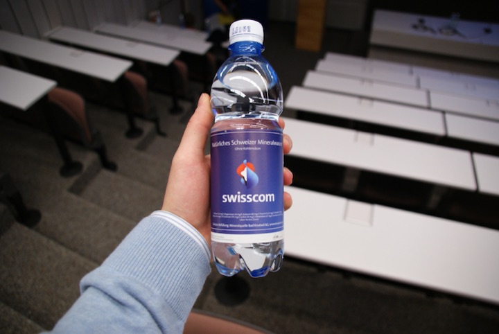 Hand holding a Swisscom mineral-water bottle in a classroom.