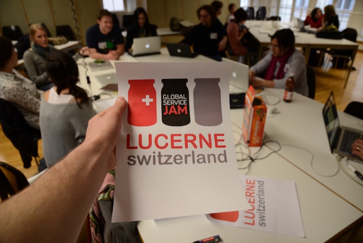 Hand holding a Global Service Jam Lucerne sign in a workshop room with participants at tables.