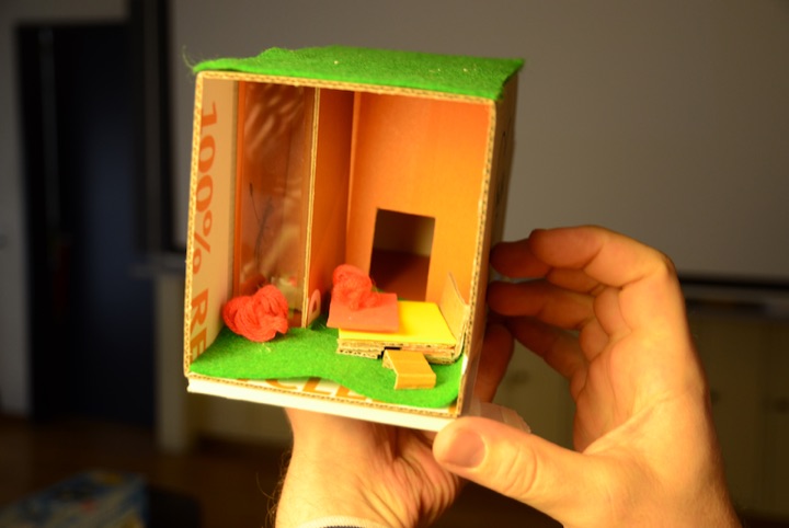 Hand holding a small cardboard room model with a green floor and orange walls.