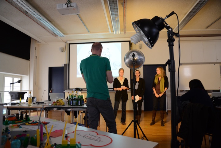 Workshop participants presenting at the front of a room beside studio lights and prototypes.