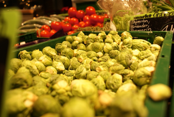 Crate of Brussels sprouts in a market display with tomatoes behind it.