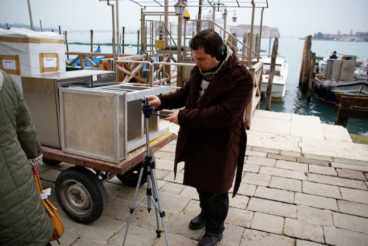 Person adjusting a tripod beside a handcart on a waterfront quay.