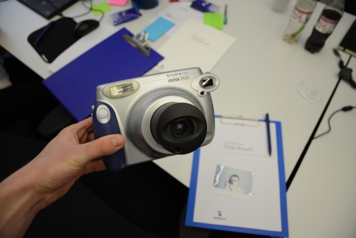 Hand holding an instant camera above a tabletop with a clipboard, notes, and a small photograph.