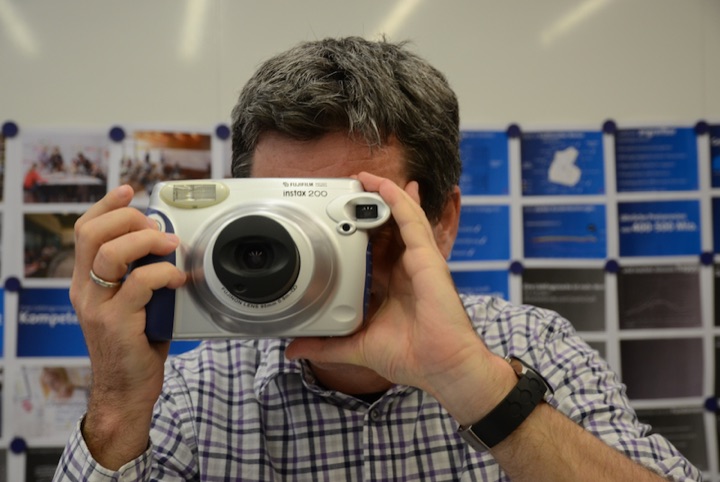 Person holding a large instant camera in front of a board of pinned photographs and blue cards.