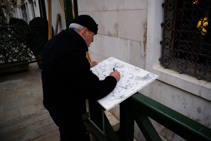 Person in a black coat and cap drawing at an outdoor stand.