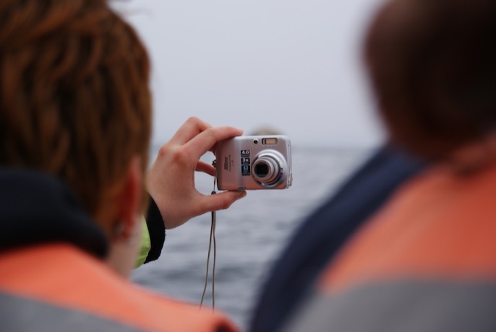 Person holding a compact camera in front of the sea, seen between two out-of-focus people.