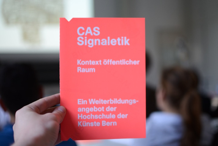 Hand holding a red CAS Signaletik flyer in front of seated people.