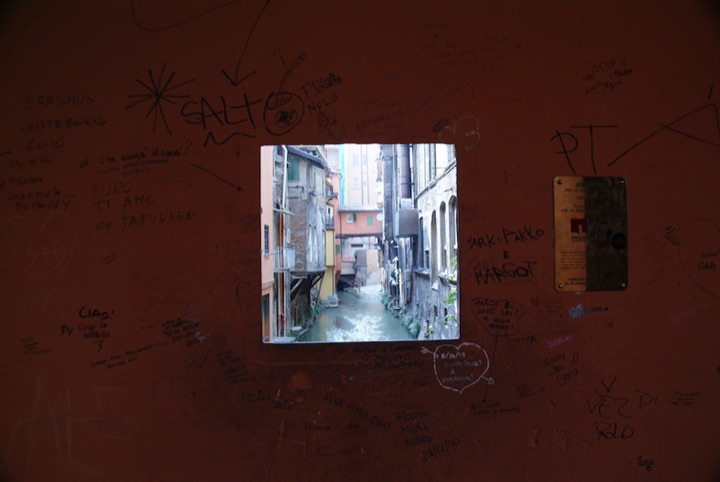 Rectangular opening in a graffiti-covered wall framing a view of buildings along a narrow canal.