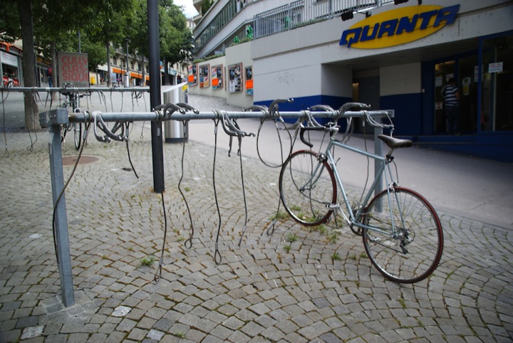 One bicycle locked to a metal rail amid empty locks at a bicycle stand outside a store.