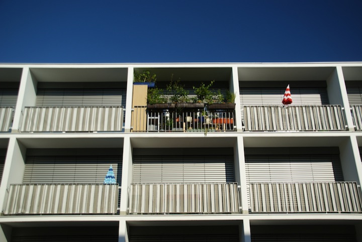 Apartment building facade with balconies, potted plants, and striped awnings against a blue sky.
