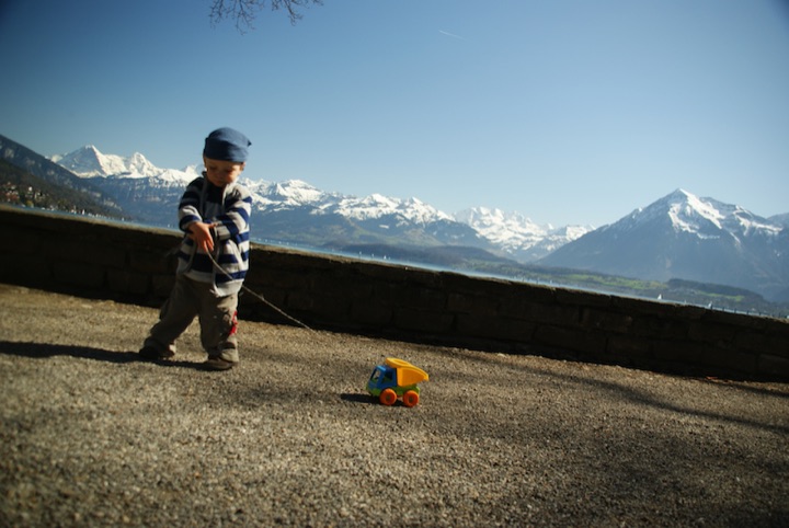 Young child pulling a small toy dump truck with a lake and snow-covered mountains in the background.
