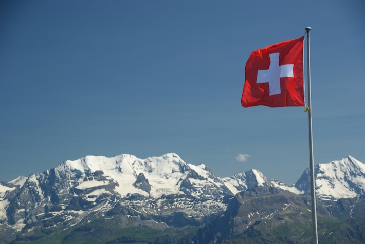 Swiss flag flying on a pole in front of snow-covered mountains under a clear blue sky.