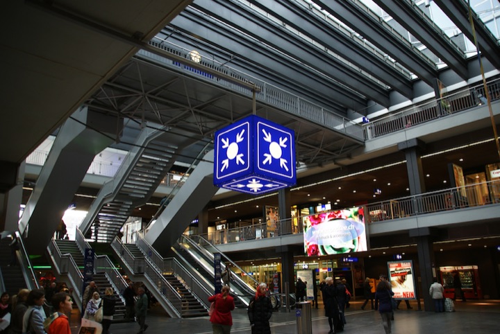 Blue meeting-point sign hanging above escalators in a multi-level station concourse.