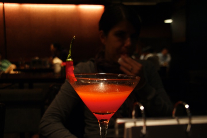 Red cocktail in a martini glass with a green garnish, reflected beside a seated person in a dim restaurant.