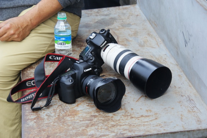 Two Canon cameras with lenses resting on a concrete bench beside a seated person and a water bottle.
