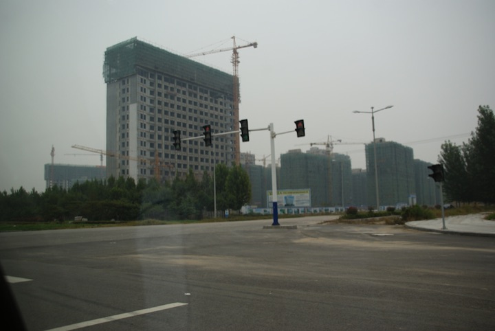 High-rise buildings under construction with cranes beside a wide road and traffic lights in hazy weather.