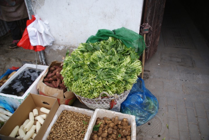 Basket of leafy green vegetables beside boxes of corn, peanuts, and sweet potatoes at a street market.