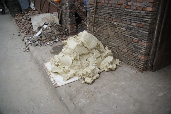Pile of pale yellow fibrous material on a pavement beside a weathered brick wall.