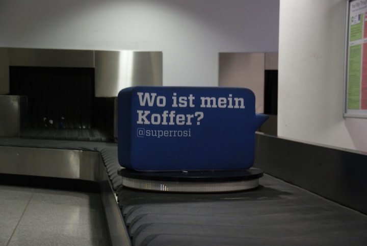 Blue suitcase-shaped advertisement reading ‘Wo ist mein Koffer? @superrosi’ on an airport baggage carousel.
