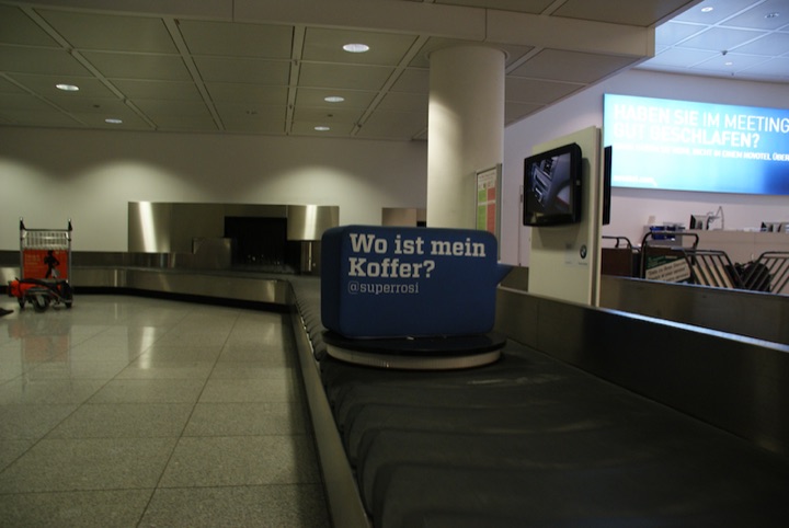 Airport baggage carousel with an advertising-covered suitcase and digital advertisements in the background.