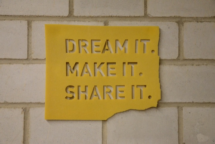 Yellow plaque reading ‘DREAM IT. MAKE IT. SHARE IT.’ mounted on a white brick wall.