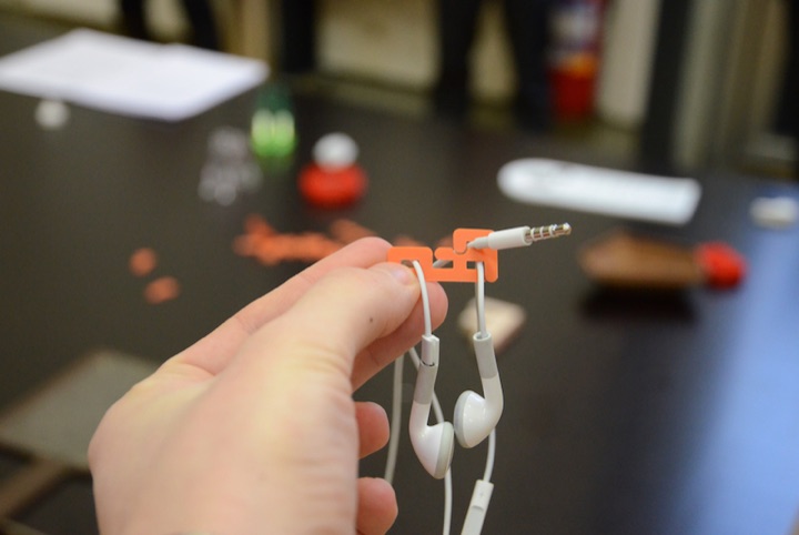 Hand holding a small orange clip that organizes a pair of white earbuds.