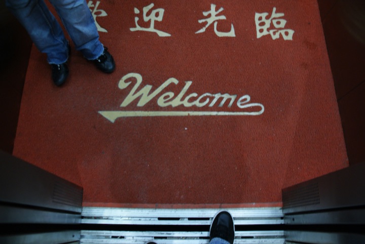 Red entrance mat with Chinese characters and the word “Welcome,” viewed from an open doorway.