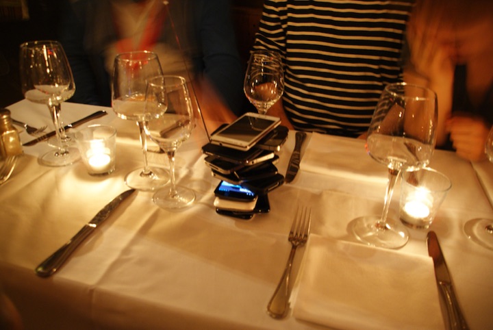 Stack of mobile phones at the center of a restaurant table set with glasses, candles, and cutlery.