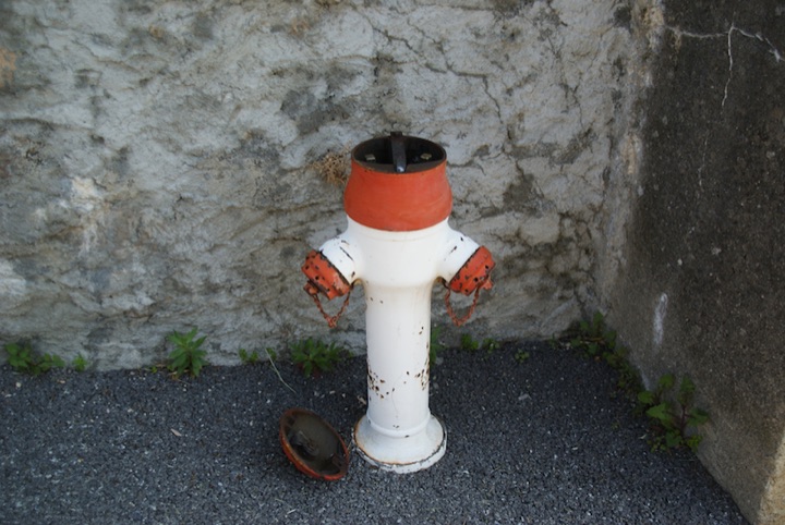 White and red fire hydrant with its top missing and a detached circular cover on gravel beside a stone wall.