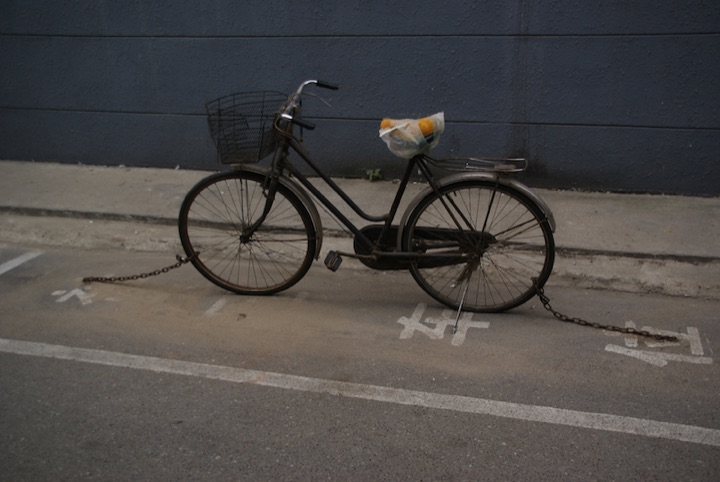 A black bicycle secured with chains in a marked parking space against a dark wall.